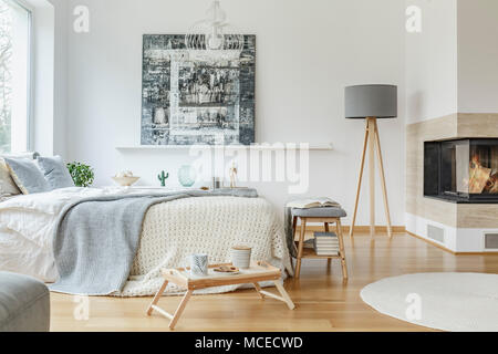 Grey lamp next to a fireplace in bright bedroom interior with painting on the wall and blanket on bed Stock Photo