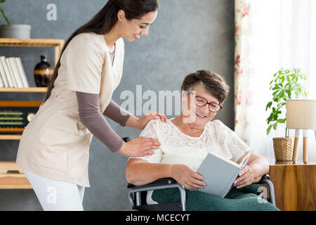 Young nurse taking care of a happy senior woman in a wheelchair who is reading a book Stock Photo