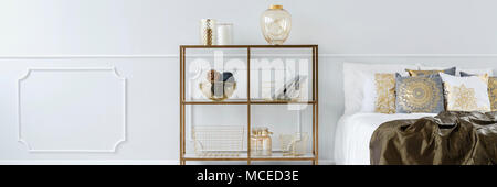 Gold rack with books and decorations standing next to bed with many cushions in bright bedroom interior Stock Photo