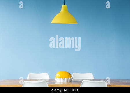 Minimal style, symmetric interior with a yellow lamp hanging over a wooden table with a melon in a dish standing on it as well as visible tops of chai Stock Photo