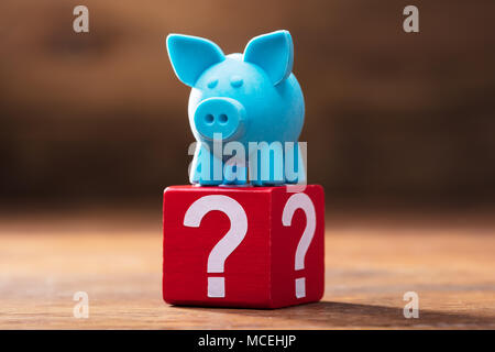 Close-up Of Blue Piggybank On Red Block With Question Marks Over The Wooden Desk Stock Photo