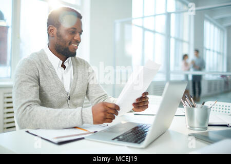 Waist-up portrait of cheerful African American businessman studying document while sitting at desk of modern open plan office, lens flare Stock Photo