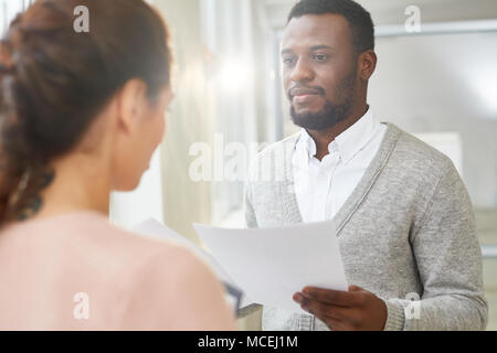 Handsome African American superior holding documents in hands while analyzing results of accomplished work with young subordinate, lens flare Stock Photo