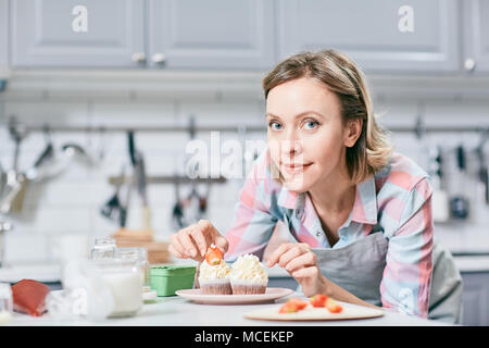Portrait of attractive Caucasian woman decorating appetizing cupcakes with strawberries and smiling at camera Stock Photo