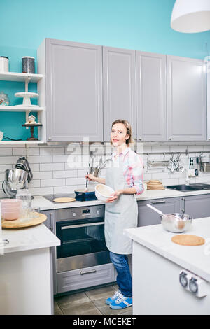 Full-length portrait of Caucasian woman standing by electric range, preparing meal in frying pan and looking at camera Stock Photo