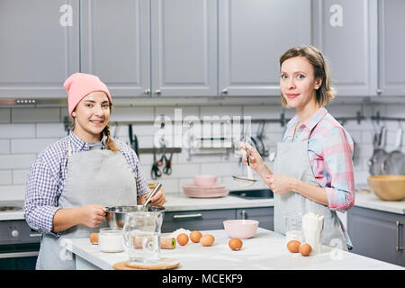 Portrait of young attractive female cooks smiling at camera while whisking eggs and sifting flour for cake in kitchen Stock Photo