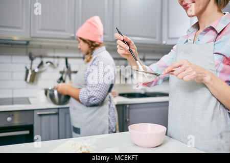 Smiling Caucasian woman sifting flour through sieve while her female colleague whisking eggs in blurred background Stock Photo