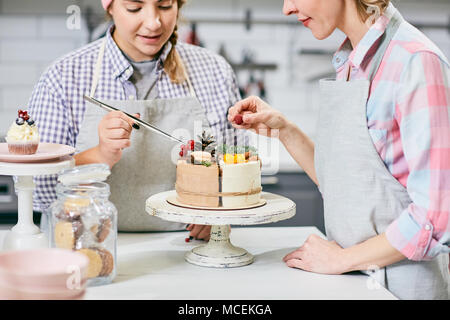 Young Caucasian female confectioners decorating delicious cake with cones, fir twigs and fresh berries in kitchen Stock Photo