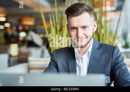 Portrait of young attractive Caucasian man in business suit sitting at laptop in cafe and smiling at camera while waiting for lunch Stock Photo
