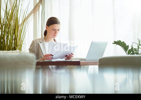 Young attractive Caucasian woman in formal wear looking through documents while waiting for lunch in cafe Stock Photo
