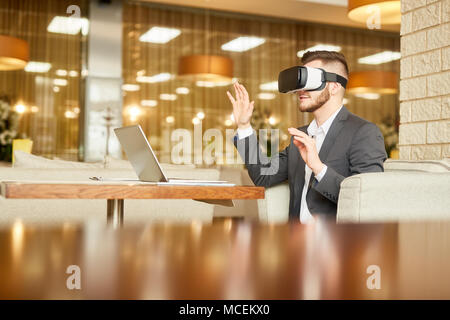 Young Caucasian manager in head-mounted display having fun while sitting at cafe table with laptop and waiting for lunch Stock Photo