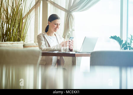 Young attractive woman in elegant business suit texting on smartphone and smiling while sitting in cafe on lunch break Stock Photo