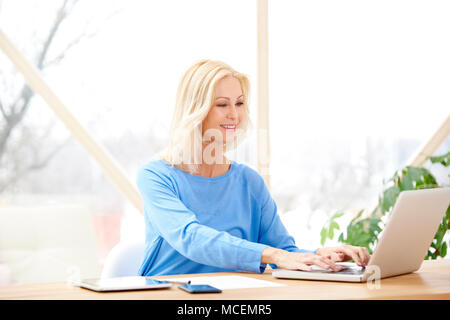 Smiling middle aged businesswoman typing on laptop while sitting at desk and working from home. Home office. Stock Photo