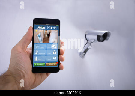 Human Hand Using Smart Home Application On Smartphone Near CCTV Camera Mounted On Wall Stock Photo
