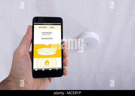 Close-up Of A Human Hand Holding Mobile Phone In Front Of Smoke Detector On Ceiling Stock Photo