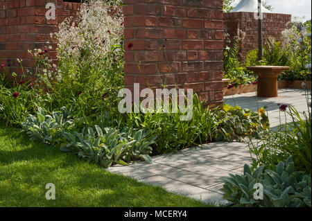 Sunny patio seating area & flowering border plants seen through gaps in walls in 'The Albert Dock Garden' - RHS Flower Show, Tatton Park, England, UK. Stock Photo