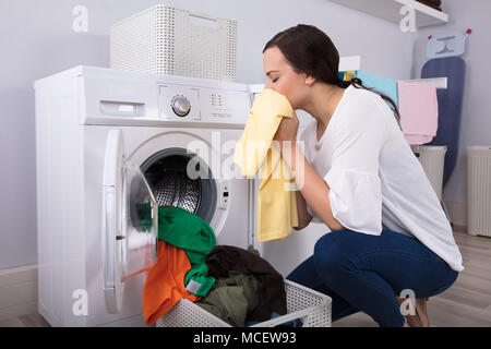 Side View Of A Young Woman Smelling Cleaned Yellow Cloth Near Washing Machine Stock Photo