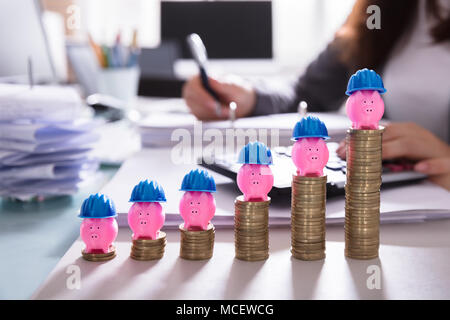 Close-up Of Pink Piggybank With Blue Hard Hat Over Increasing Stacked Coins At Workplace Stock Photo