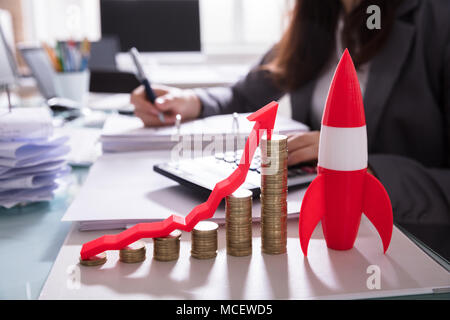 Close-up Of Red Rocket Besides Stacked Coins And Arrow Showing Upward Direction On Wooden Desk Stock Photo