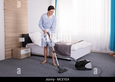 Happy Young Housekeeper Cleaning Carpet With Vacuum Cleaner In Hotel Room Stock Photo