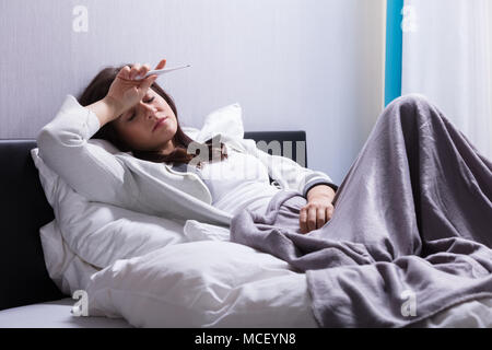 Sick Young Woman Lying On Bed Holding Thermometer In Bedroom Stock Photo