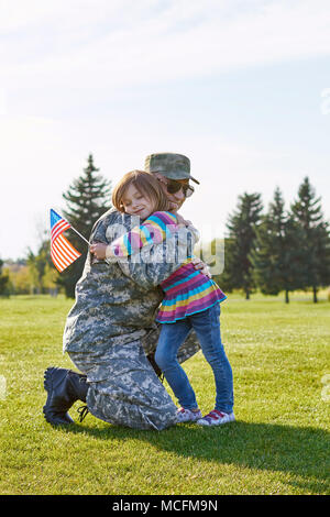 Little kid with usa flag is embracing her father. Military daddy is hugging with his little daughter on the park lawn. Stock Photo