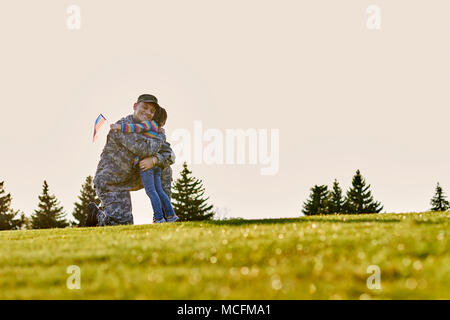 American soldier is hugging his little daughter outdoor. Reunion of father in military uniform and daughter in the park lawn. Stock Photo