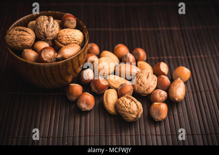 Different nuts in a wooden bowl on a dark bamboo surface. Some of them have been spilt over from the bowl. Stock Photo