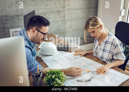 Young pensive architect colleagues analyzing blueprints and talking about him at the office. Stock Photo