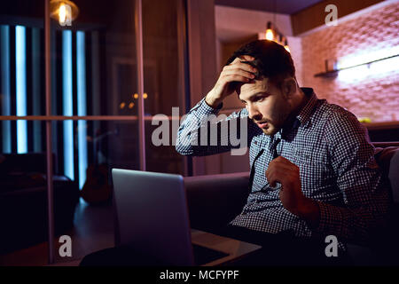 Problems, stress, depression, crisis. A young man with a laptop holds his head with his hand sitting at night in the room. Stock Photo