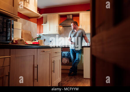 Mature man is standing in the kitchen of his home with a cup of tea in his hand, staring deeply out of the window. Stock Photo