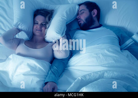 Couple in bed, man snoring and woman can't sleep, covering ears with pillow for snore noise Stock Photo