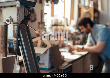 Closeup of a drill press in a woodworking workshop with a carpenter using a digital tablet at a bench in the background Stock Photo