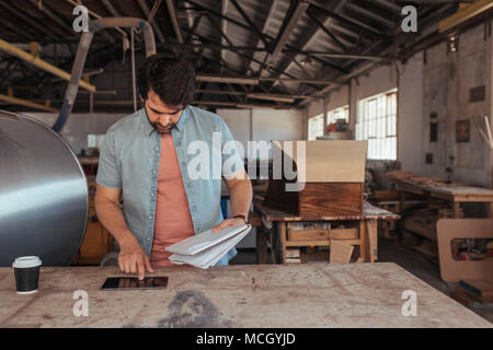 Skilled young woodworker standing at a workbench in his carpentry shop using a digital tablet and holding a notebook of designs Stock Photo