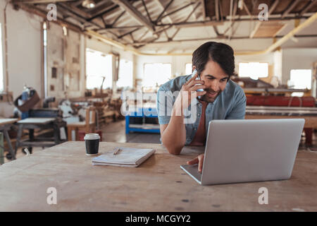 Smiling young craftsman standing at a workbench in his large workshop working online with a laptop and discussing designs with a customer on a cellpho Stock Photo