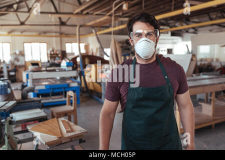 Portrait of a young craftsman wearing an apron and a protective mask and glasses working alone in his large carpentry workshop Stock Photo