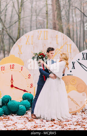 Happy wedding couple softly hugs at the big vintage clocks in  autumn forest. Creative decorations Stock Photo