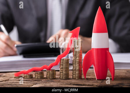 Close-up Of Red Rocket Besides Stacked Coins And Arrow Showing Upward Direction On Wooden Desk Stock Photo