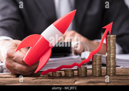 Businessperson's Hand Flying Red Rocket Over Stacked Coins And Arrow Showing Upward Direction Stock Photo