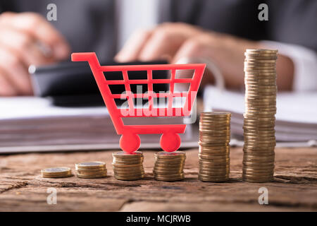 Close-up Of Red Shopping Cart On Stacked Golden Coins Over Wooden Desk Stock Photo
