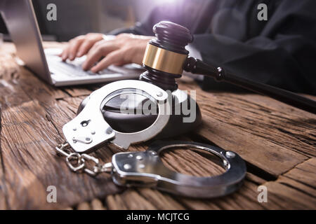 Close-up Of Handcuffs And Gavel In Front Of Judge Working On Laptop Stock Photo