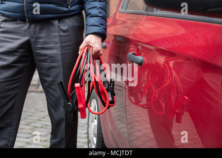 Close-up Of A Person's Hand Holding Jumper Cables Near Red Car Stock Photo