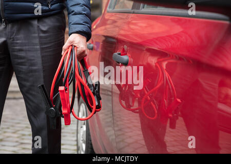 Close-up Of A Person's Hand Holding Jumper Cables Near Red Car Stock Photo