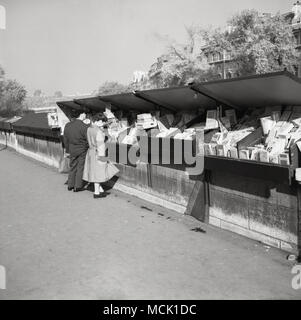 1950s, historical picture of a Parisian couple looking at the stock at a Bouquiniste in Paris, France. These independent booksellers of used and antiquarian books trade from metal boxes along the sections of the banks of the River Seine and have been a major feature of Parisian life for over 100 years. Stock Photo