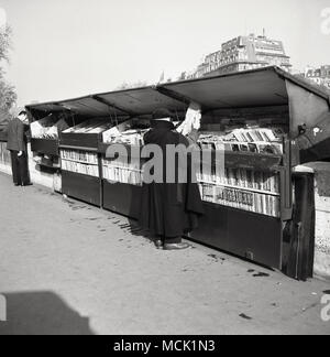 1950s, historical picture of a Parisian man wearing a beret and a long cape looking at the stock at a Bouquiniste in Paris, France. These independent booksellers of used and antiquarian books trade from metal boxes along the sections of the banks of the River Seine and have been a major feature of Parisian life for over 100 years. Stock Photo