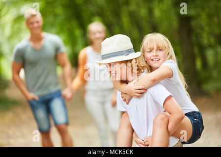 Girl is sitting on the back of her brother with parents in the background Stock Photo