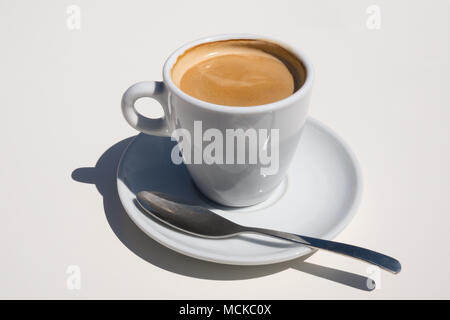 A delicious cup of hot coffee in a white cup and saucer with spoon sitting on a white table from the front. Stock Photo