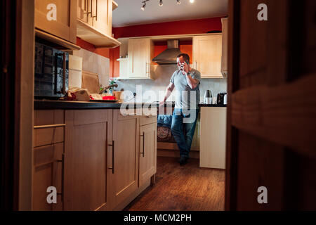 Mature man is standing in the kitchen of his home, looking out the window while talking to someone on his smartphone. Stock Photo
