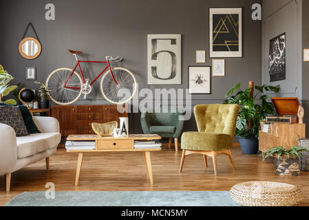 Art collection on a grey wall, wooden coffee table, armchair and red bike in a messy living room interior Stock Photo
