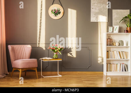 Pink armchair next to a table with red tulips in cozy grey living room interior with decor on the wall Stock Photo